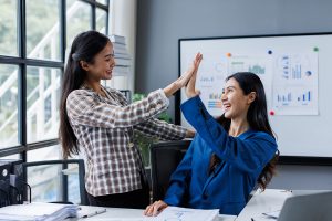 Two women high-five in an office room full of charts and graphs